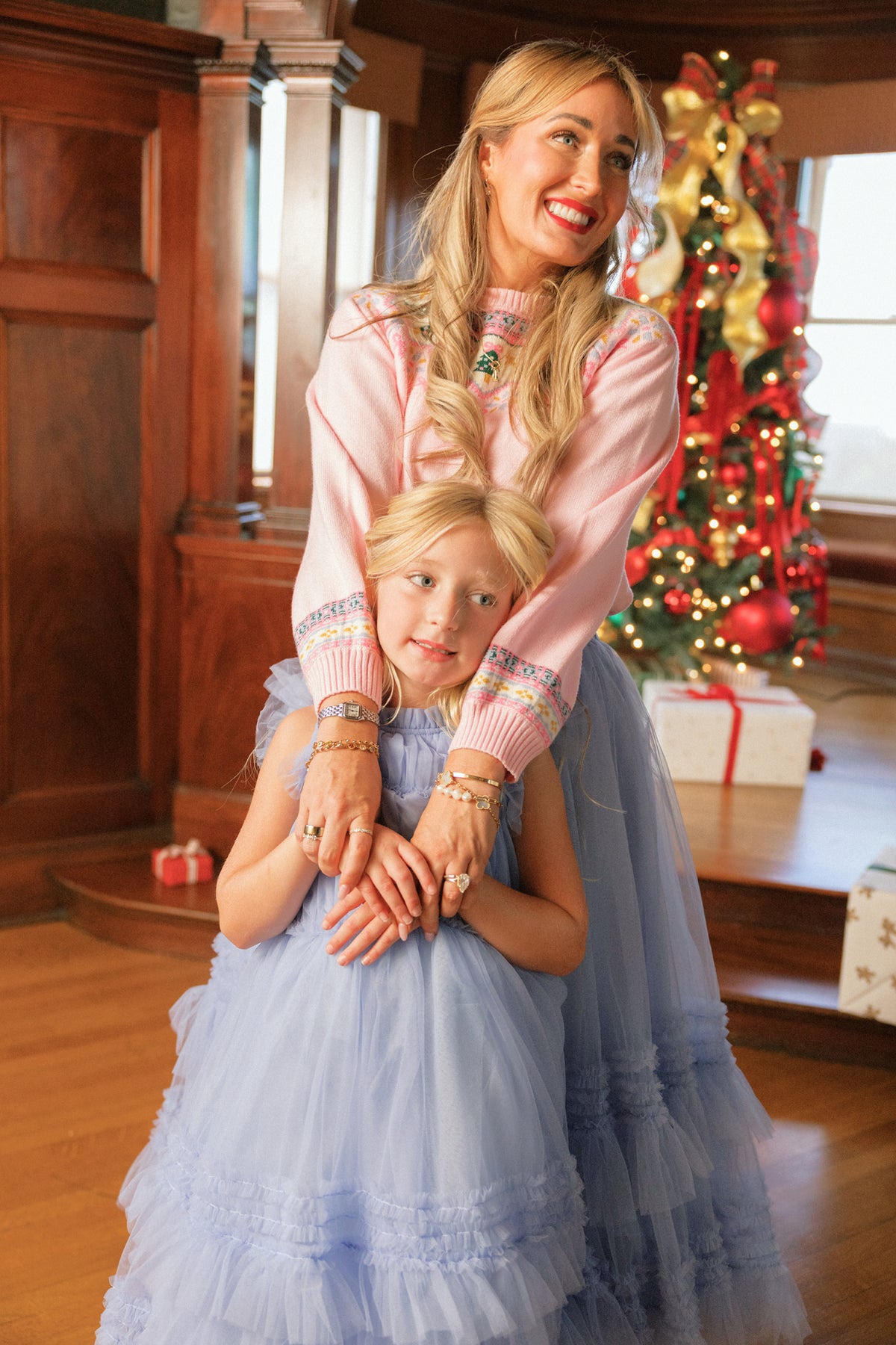 A smiling woman in a pink top hugs a young girl wearing the Ivy City Co Celestine Skirt in Misty Blue, in front of a decorated Christmas tree with wrapped presents underneath.