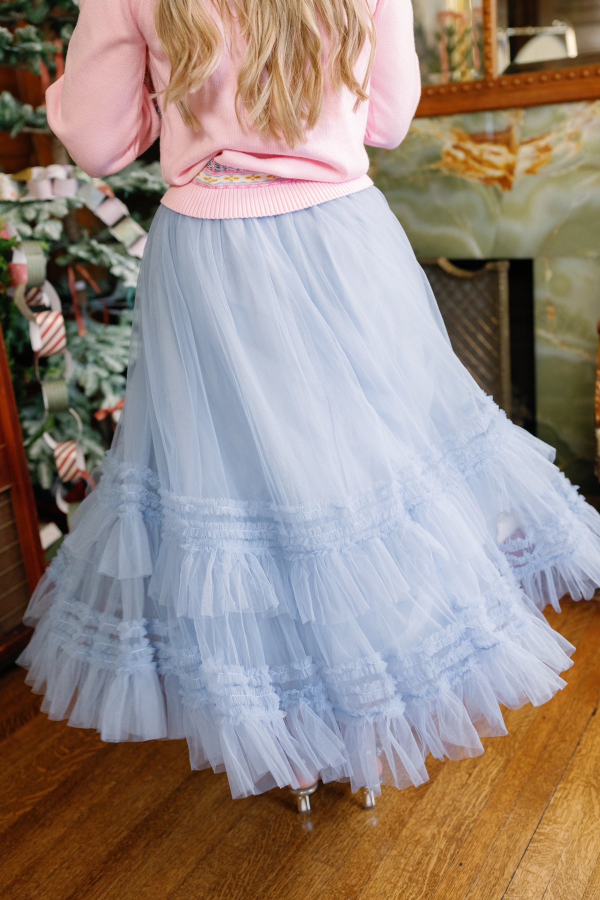 A person wearing the Ivy City Co Celestine Skirt in Misty Blue and a pink sweater stands indoors near a decorated Christmas tree on wood flooring, creating the perfect holiday recital outfit.