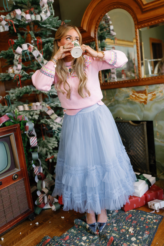 Wearing the Ivy City Co Celestine Skirt in Misty Blue and a pink sweater, a woman sips from a mug beside a decorated Christmas tree, surrounded by gifts and a vintage TV—creating the perfect holiday recital look.