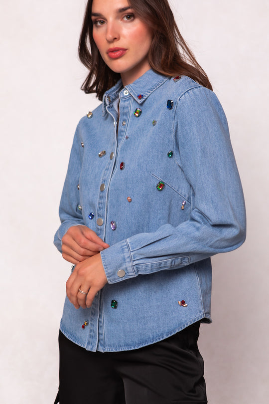 A woman with long brown hair wears Ivy City Co’s Rhinestone Cowboy Shirt, a light blue denim top with colorful rhinestones, paired with black pants. She poses with one hand near her face and the other at her side.