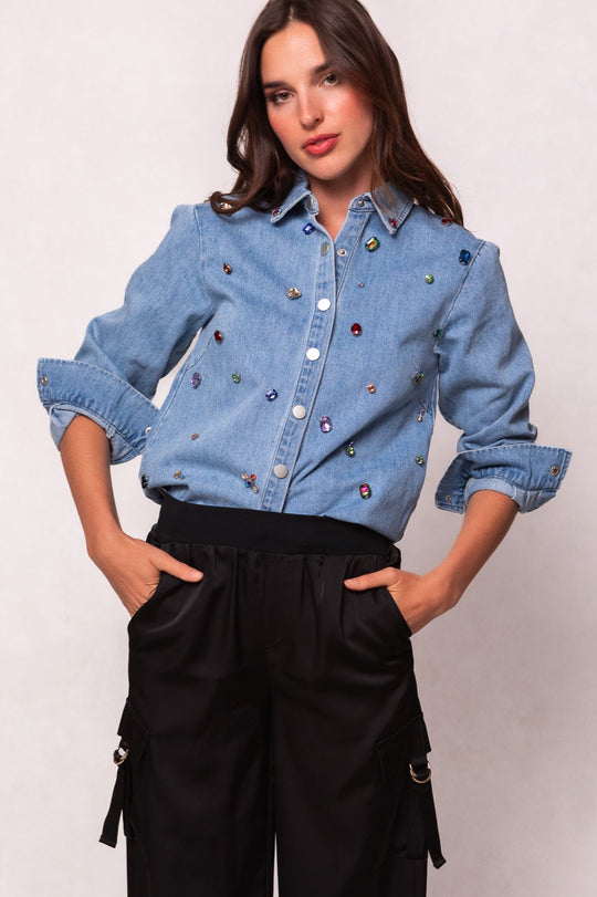 A woman with long dark hair and a neutral expression stands with her hands in her black cargo pant pockets, wearing the Ivy City Co Rhinestone Cowboy Shirt in light blue, featuring colorful jewel embellishments.