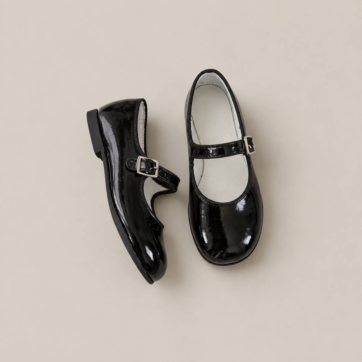 The Mini Mary Janes in Black Patent by Zimmerman Shoes, featuring adjustable ankle straps and buckled closures, are displayed on a neutral beige background.