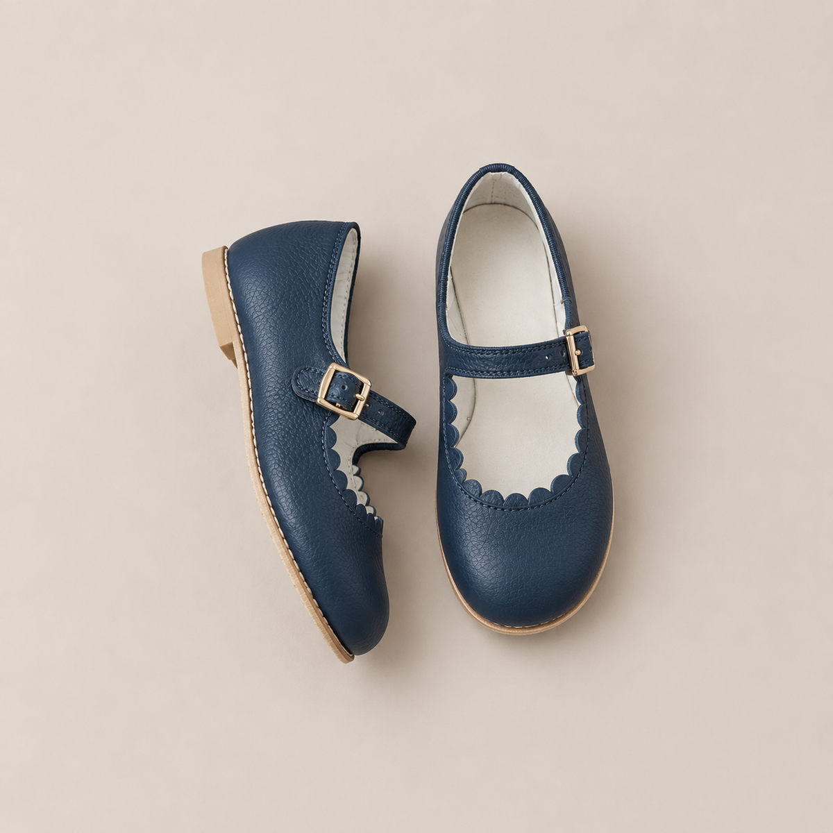 The Mini Mary Janes in Scalloped Navy by Zimmerman Shoes are crafted from navy leather with gold buckles and an adjustable ankle strap, and are showcased on a light beige background.