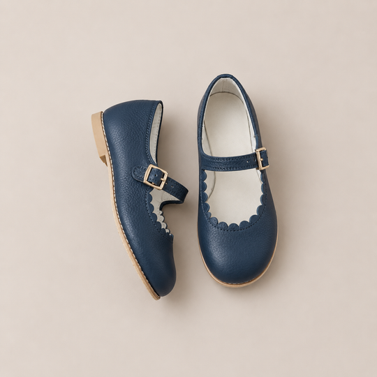 The Mini Mary Janes in Scalloped Navy by Zimmerman Shoes are crafted from navy leather with gold buckles and an adjustable ankle strap, and are showcased on a light beige background.