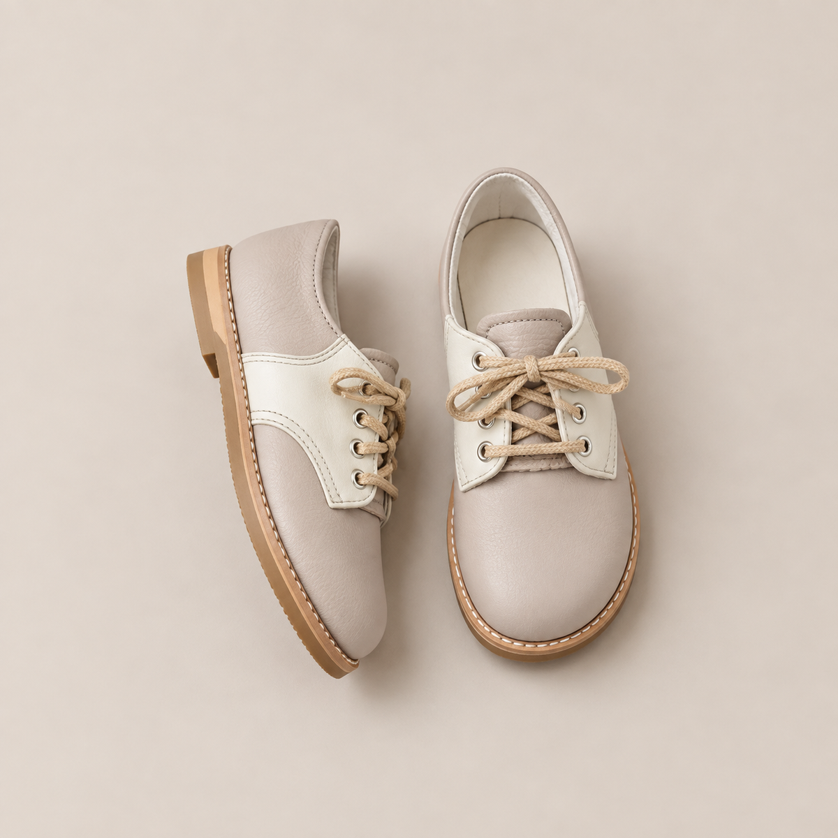 Two Mini Boys Saddles in Sand & Fog by Zimmerman Shoes—beige leather, oxford-style children’s shoes with tan rubber soles—are placed on a light, neutral background. Reminiscent of classic saddle shoes, one shoe is upright while the other lies on its side, showcasing the laces and detailed stitching characteristic of formal footwear.