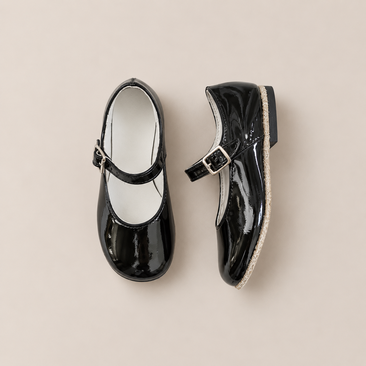 A pair of Zimmerman Shoes’ Mini Mary Janes in Black Patent is displayed on a light beige background—one upright and the other on its side—highlighting their glossy finish, flat sole, and buckled adjustable ankle straps.