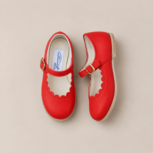 A pair of Mini Mary Janes in Scalloped Red by Zimmerman Shoes, featuring bright red leather with scalloped edges and adjustable ankle straps, displayed on a light beige background.