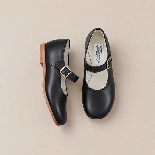 Two black Mini Mary Janes from Zimmerman Shoes with tan soles and gold buckles rest on a beige background—one shoe upright, the other lying on its side to reveal the strap and interior logo. These classic children’s dress shoes blend timeless style with comfort, making them perfect for any special occasion.