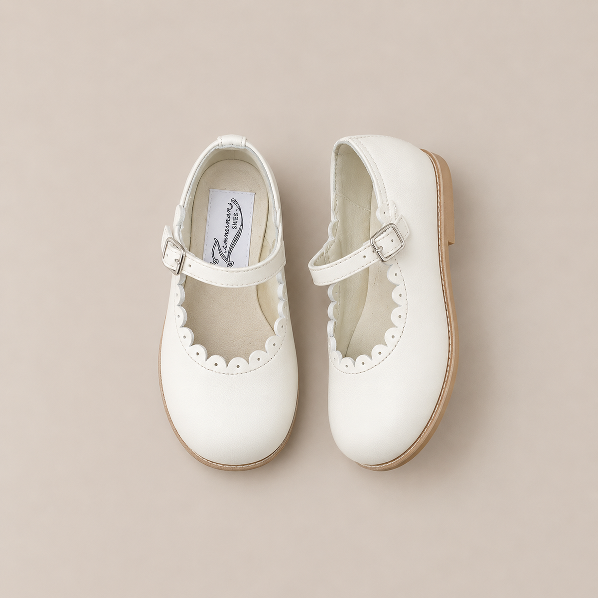 A pair of Zimmerman Shoes Mini Mary Janes in Scalloped Pearl, crafted from off-white leather with adjustable ankle straps, delicate scalloped edges, and light brown soles, displayed on a neutral beige background.
