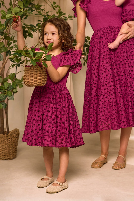 A young girl wearing the Ivy City Co Mini Hattie Dress in Magenta stands beside an adult in a matching dress, holding a basket and touching a leafy indoor plant. The legs of another small child, held by the adult, are also visible.