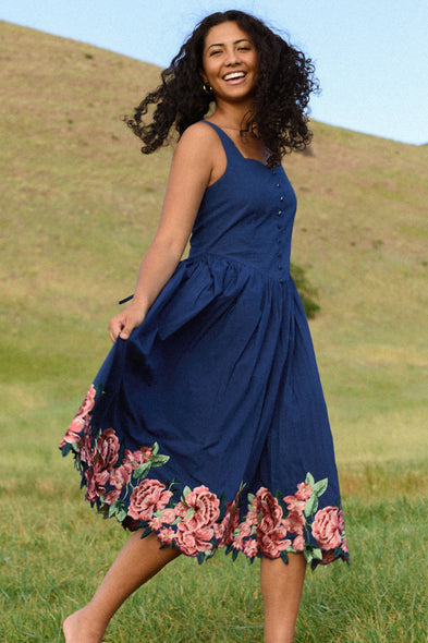 A smiling woman in the Ivy City Co Clarissa Dress in Midnight with Red Flowers twirls outdoors on a grassy hillside under a clear sky.