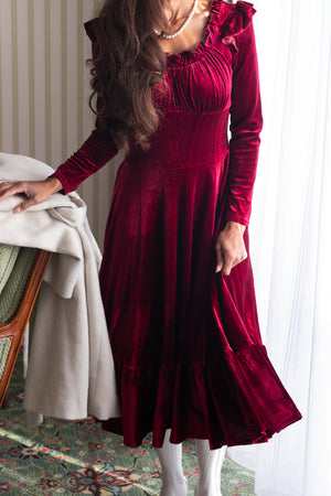 A person wearing a wine red velvet dress with a sweetheart neckline and long sleeves. The dress features a fitted waist and a midi-length skirt, with a visible hidden back zipper.