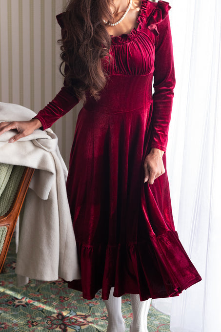 A person wearing a wine red velvet dress with a sweetheart neckline and long sleeves. The dress features a fitted waist and a midi-length skirt, with a visible hidden back zipper.