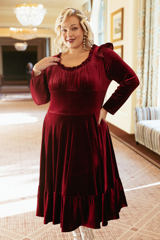 A person wearing a red wine velvet dress with a sweetheart neckline and long sleeves, standing next to a chair in a room with light decor.