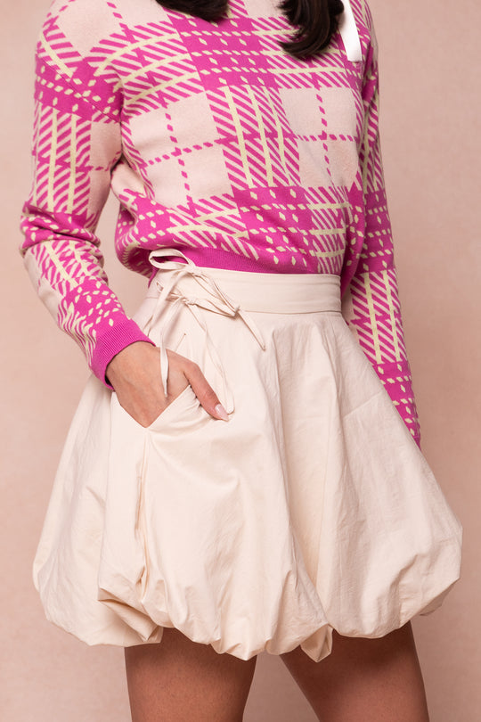 A woman wears the Ivy City Co Clo Bubble Skirt in Cream with a pink and cream plaid sweater. Her hand is in the skirt pocket, facing away from the camera against a soft beige background. Her face is not visible.
