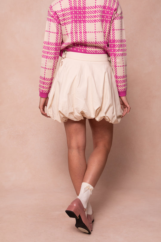 A person faces away, wearing the Ivy City Co Clo Bubble Skirt in Cream, paired with a pink and white plaid sweater, white socks, and pink shoes against a beige background.