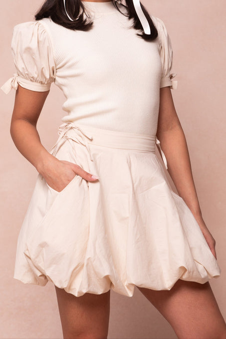 A person wears the Ivy City Co Clo Bubble Skirt in Cream with side bows and pockets, paired with a matching short-sleeved cream top, standing against a light beige background.