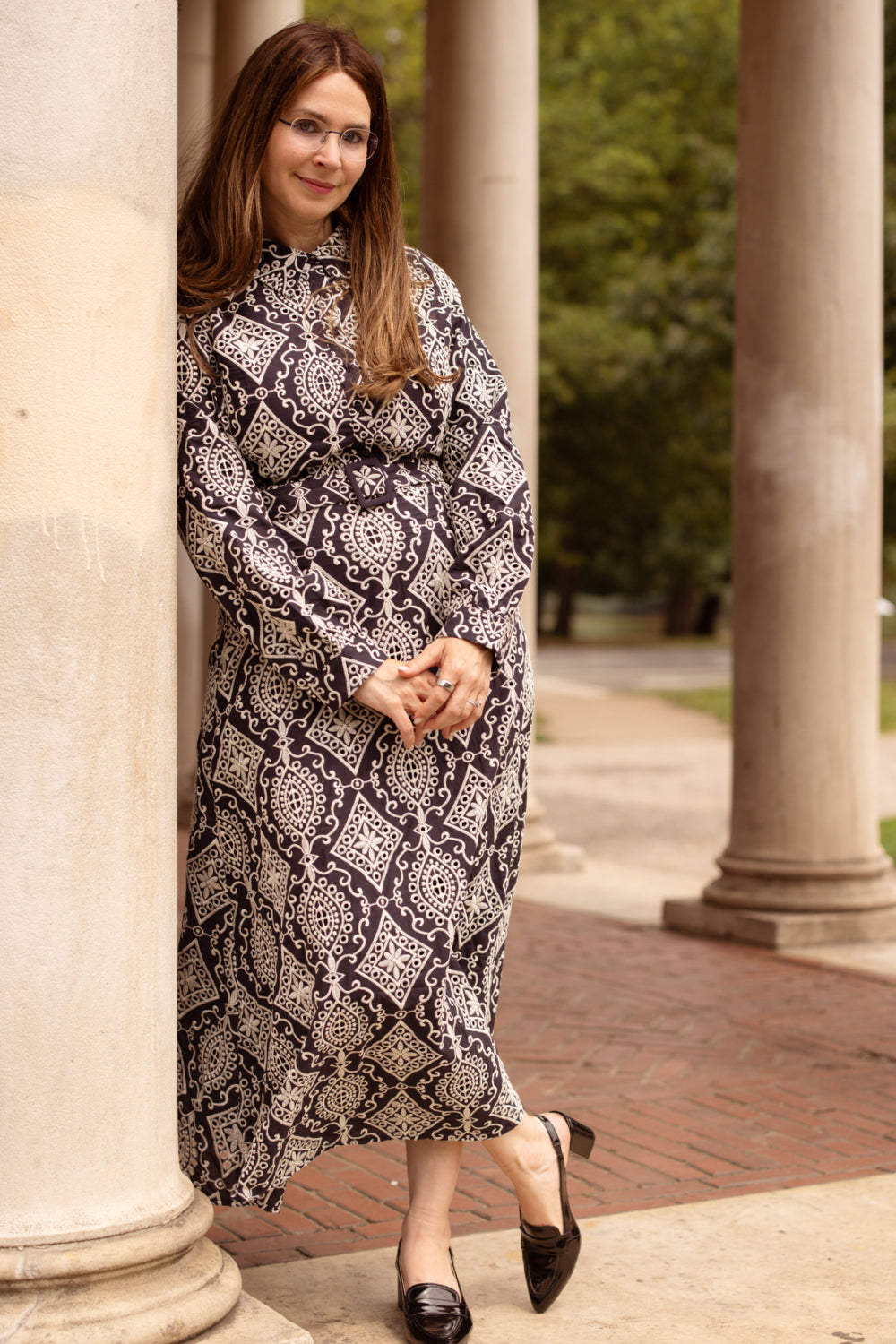 A woman wearing the Clover Dress in Midnight Tile by Ivy City Co and glasses leans against a column outdoors, smiling slightly, with trees and more columns in the background.