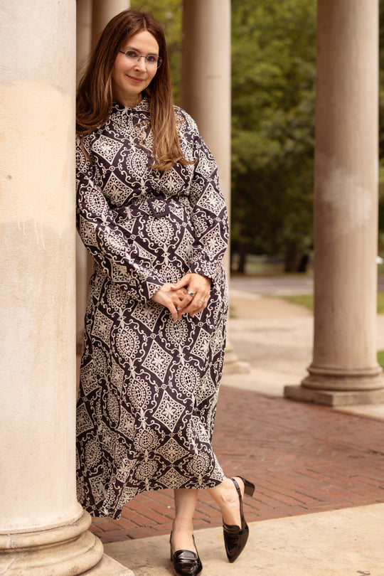 A woman wearing the Clover Dress in Midnight Tile by Ivy City Co and glasses leans against a column outdoors, smiling slightly, with trees and more columns in the background.
