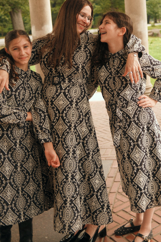 Three girls stand close together, smiling and wearing matching Ivy City Co Clover Dresses in the Midnight Tile print—a striking black-and-white pattern. These ankle-length dresses create a coordinated look as they embrace, radiating happiness beneath a covered outdoor area. With their nursing friendly design, these Clover Dresses offer both style and comfort for any occasion.