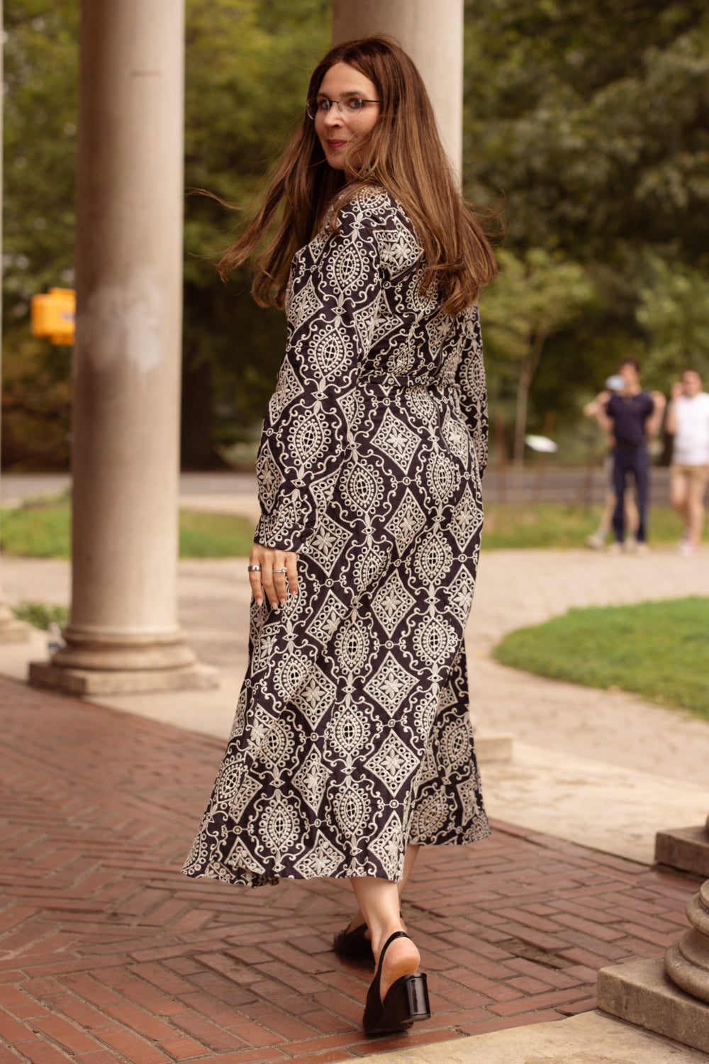 A woman in the Ivy City Co Clover Dress in Midnight Tile and heeled sandals walks away, glancing back over her shoulder. She is outdoors near columns and greenery, with a few people visible in the background.