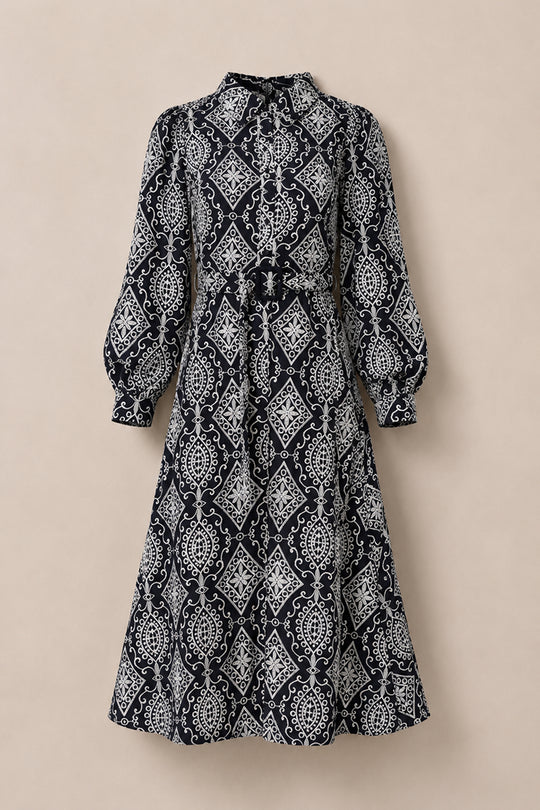 The Clover Dress in Midnight Tile by Ivy City Co features long sleeves, a midi length, and a high collar with intricate white geometric and floral patterns set against a deep, dark background. Displayed against a neutral backdrop, this nursing-friendly dress seamlessly blends elegance with practicality, making it ideal for any occasion.