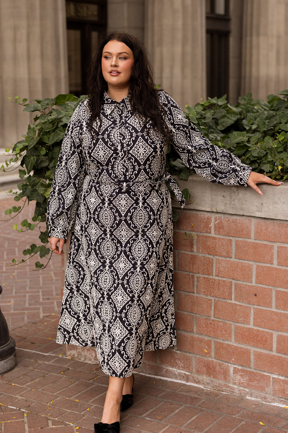 A woman in the Ivy City Co Clover Dress in Midnight Tile stands outdoors, leaning on a brick wall with greenery behind her, looking confidently to the side.