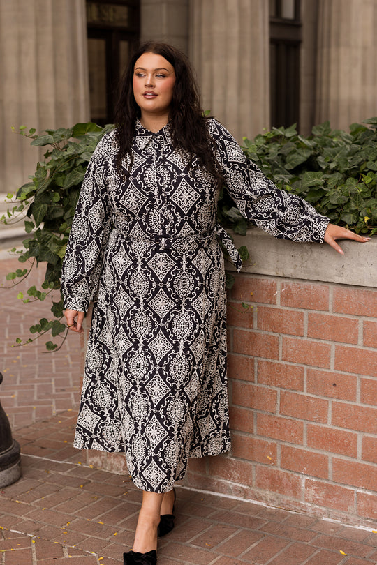 A woman in the Ivy City Co Clover Dress in Midnight Tile stands outdoors, leaning on a brick wall with greenery behind her, looking confidently to the side.