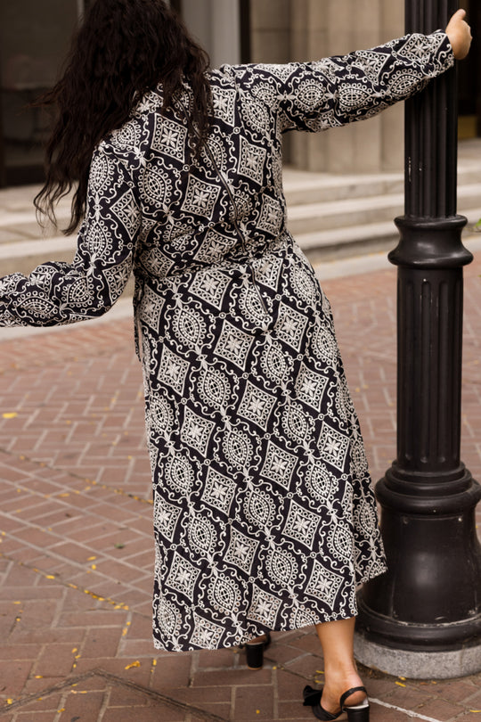 Wearing the Ivy City Co Clover Dress in Midnight Tile, a person leans back while holding a black lamppost on a brick sidewalk, facing away from the camera.