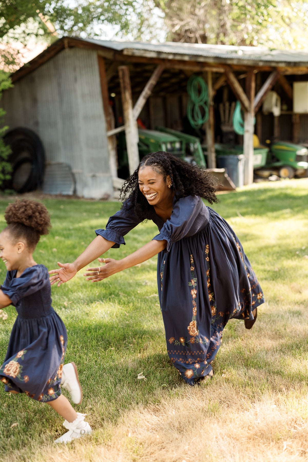 A woman and a young girl, both wearing the Ivy City Co Mini Teagan Dress in Navy Embroidered Fall Floral—with balloon sleeves and smocked bodices—play and run on the grass near a rustic shed surrounded by farm equipment. The woman smiles and reaches out as the girl runs ahead of her.