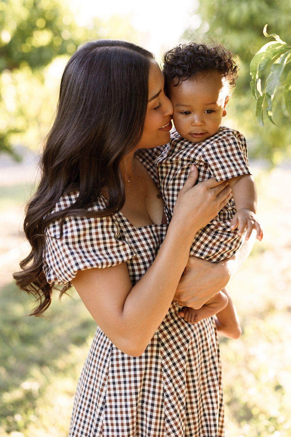 A woman stands outdoors, holding a small child as sunlight filters through green trees. Both wear matching brown and white gingham outfits from Ivy City Co—the woman in the Connie Short Sleeve Dress in Chestnut Gingham, and the child in a coordinating set. Smiling, the woman kisses the child’s cheek, their outfits perfectly paired for a sunny day.