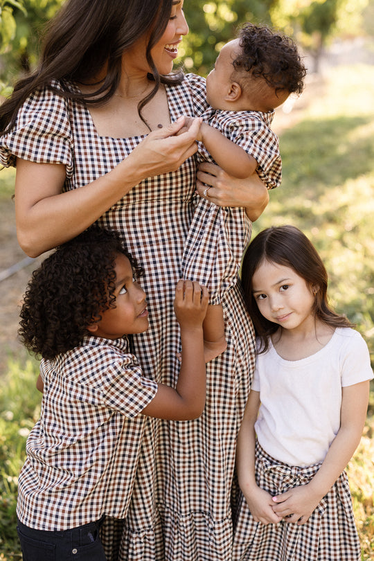 A woman stands outdoors with two children, all smiling and surrounded by greenery. They are dressed in coordinated checkered outfits from Ivy City Co, including the charming Connie Short Sleeve Dress in Chestnut Gingham. The girl, however, wears a white shirt paired with a matching chestnut gingham skirt.