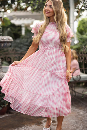 A woman with long blonde hair wears the Ivy City Co Cordelia Dress in Desert Rose Gingham, holding out the skirt and smiling slightly on a patio with greenery. The bright image showcases this maternity-friendly dress.
