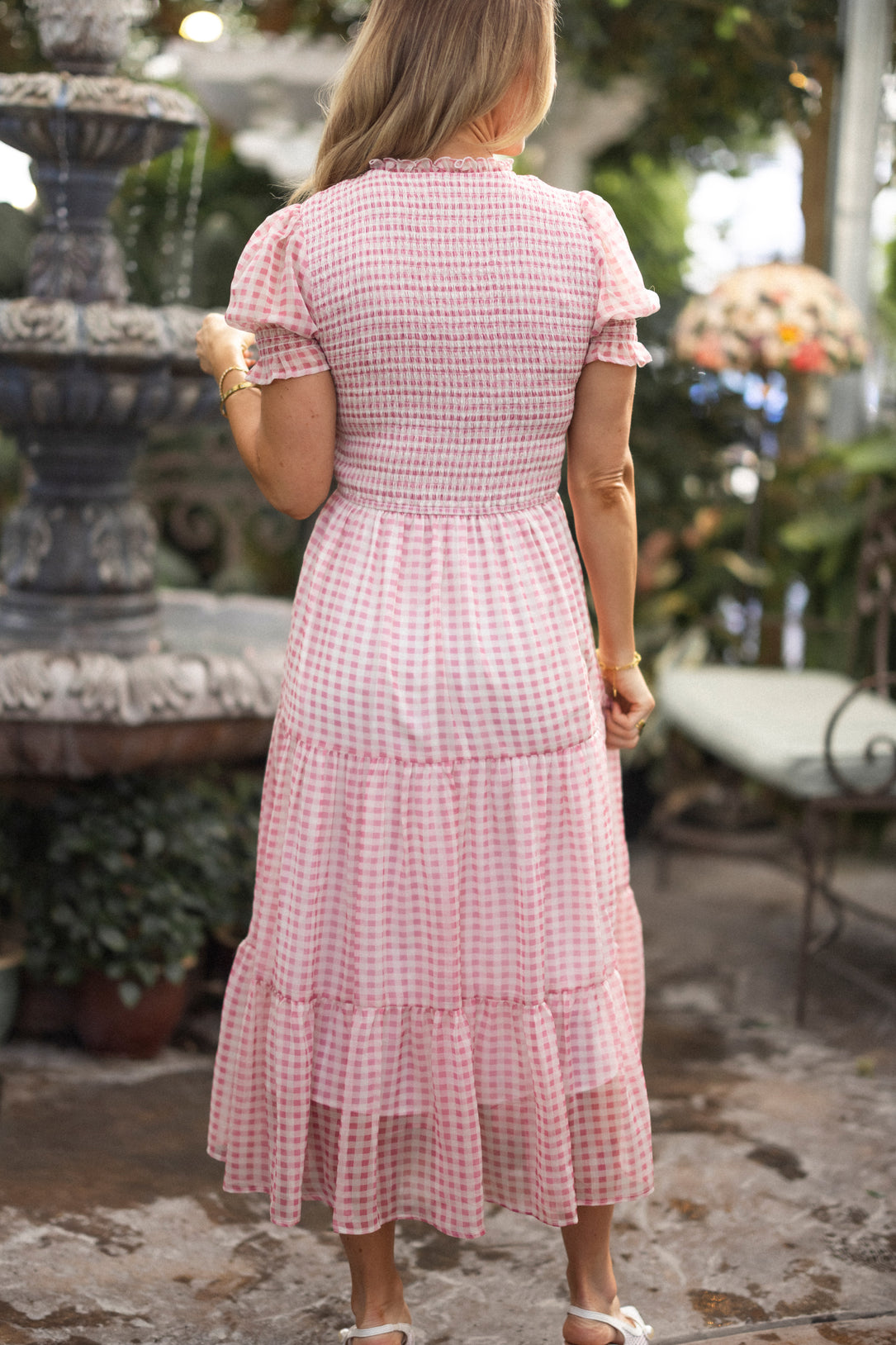 A woman with long blonde hair stands outdoors facing away, wearing the Ivy City Co Cordelia Dress in Desert Rose Gingham—maternity friendly with short puffed sleeves and ruffled tiers—by a stone fountain amid lush greenery.