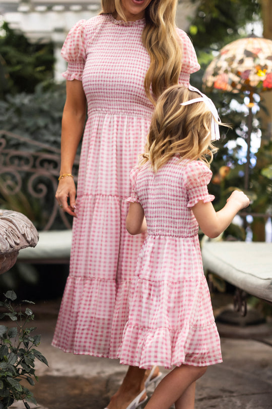 A woman and girl stand outside in matching Ivy City Co Cordelia Dresses in Desert Rose Gingham, featuring short sleeves and tiered skirts. The maternity-friendly outfits match, and the girl has a white hair bow. Their faces aren’t fully visible.