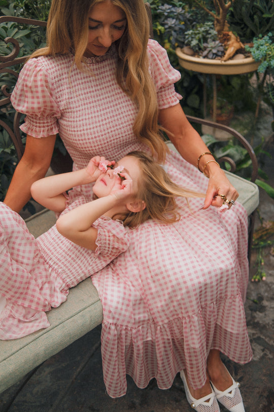 A woman and a young girl sit on a garden bench in matching Ivy City Co Cordelia Dresses in Desert Rose Gingham. The girl reclines on the woman's lap with flowers, capturing a sweet, maternity-friendly moment amid blooming plants.
