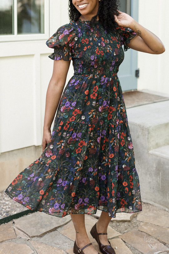 A woman wearing the Ivy City Co Cordelia Dress in Evergreen Vivid Meadow—a maternity-friendly, dark floral puff sleeve dress—stands smiling near stone steps outside a house, stylishly paired with brown shoes.