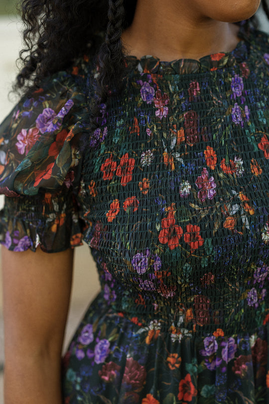A woman wearing the Cordelia Dress in Evergreen Vivid Meadow by Ivy City Co—a dark floral puff sleeve dress with red, purple, and green patterns. The smocked chest makes it maternity friendly. Her curly hair is partially visible.