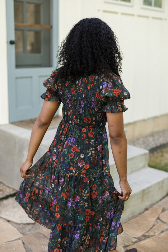A woman with curly black hair stands with her back to the camera, wearing the Cordelia Dress in Evergreen Vivid Meadow by Ivy City Co—a tiered floral puff sleeve dress with a smocked bodice. This elegant, maternity-friendly style is showcased outdoors as she holds the skirt of her dress, with a house entrance visible in the background.