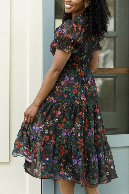 Smiling woman in the Cordelia Dress in Evergreen Vivid Meadow by Ivy City Co—a black floral puff sleeve dress adorned with red and purple flowers—stands near a blue door, facing away but turning slightly to the side. This style is both elegant and maternity friendly.