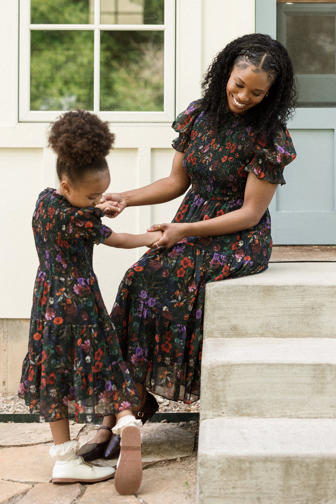 A woman and a young girl, both wearing matching Ivy City Co Cordelia Dresses in Evergreen Vivid Meadow, smile and hold hands while sitting and standing on outdoor steps near a blue door and window.