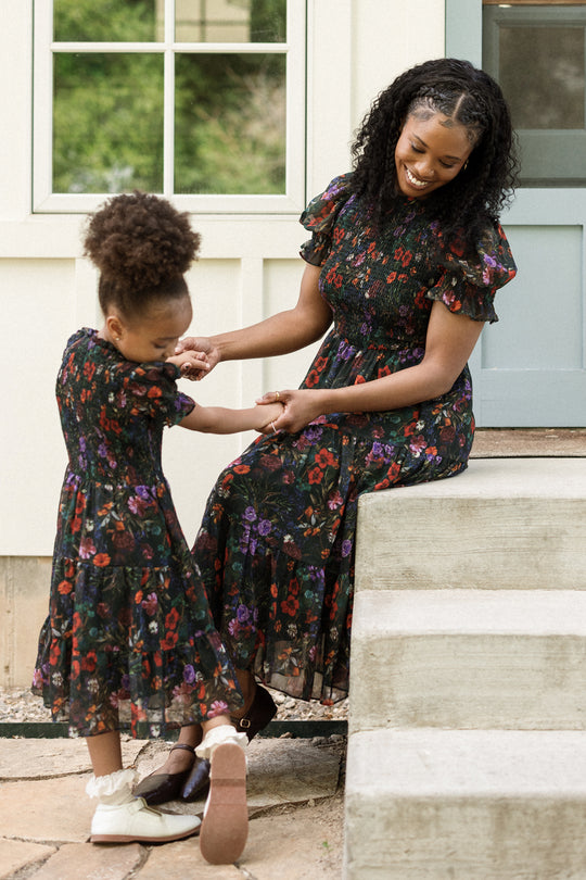 A woman and a young girl, both wearing matching Ivy City Co Cordelia Dresses in Evergreen Vivid Meadow, smile and hold hands while sitting and standing on outdoor steps near a blue door and window.