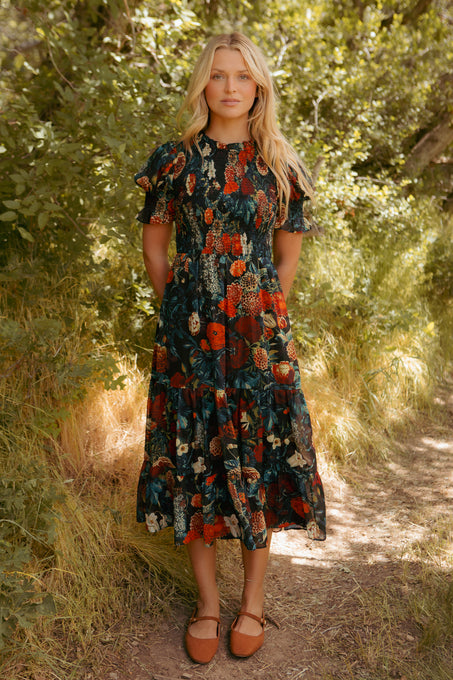 A woman with long blonde hair stands outdoors in greenery, wearing the Ivy City Co Cordelia Dress in Moody Floral—an effortlessly chic chiffon fall dress with short sleeves.
