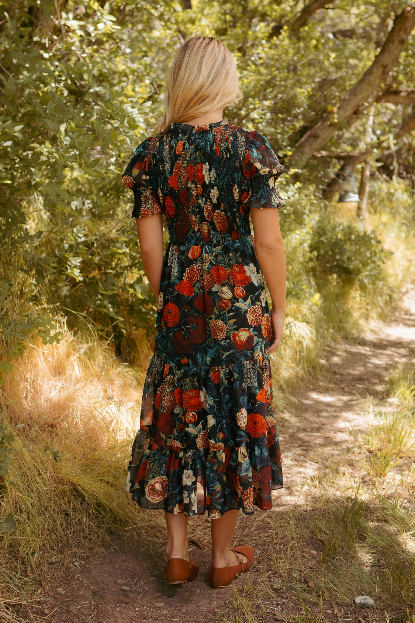 A woman with blonde hair, wearing the Ivy City Co Cordelia Dress in Moody Floral and brown shoes, stands on a sunlit forest path surrounded by greenery, facing away from the camera.
