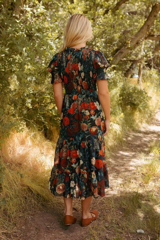 A woman with blonde hair, wearing the Ivy City Co Cordelia Dress in Moody Floral and brown shoes, stands on a sunlit forest path surrounded by greenery, facing away from the camera.