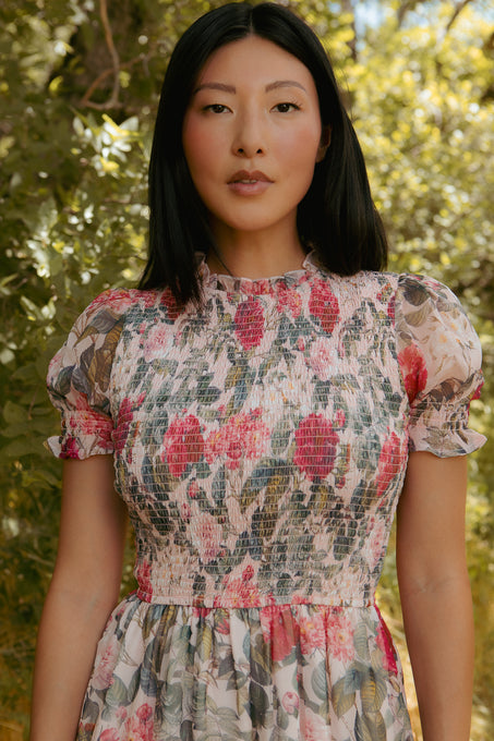 A woman with straight black hair stands outdoors in sunlight, wearing the Ivy City Co Cordelia Dress in Pink Bloom—a short-sleeved, floral chiffon midi perfect for a breezy day amidst lush greenery.