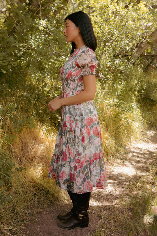 A woman with long dark hair stands sideways on a sunlit path, wearing the Ivy City Co Cordelia Dress in Pink Bloom and black boots, surrounded by green foliage and tall grass.