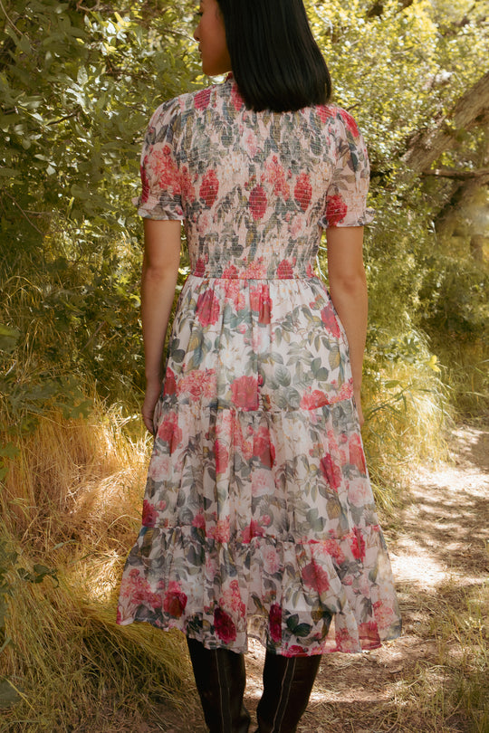 A woman with straight black hair stands on a sunlit forest path, surrounded by greenery. She wears the Ivy City Co Cordelia Dress in Pink Bloom, a light floral midi dress, paired with tall brown boots.