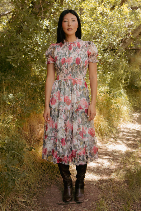 A woman stands outdoors on a dirt path, surrounded by greenery, wearing the Ivy City Co Cordelia Dress in Pink Bloom—a chic, ankle-length floral maternity dress with short sleeves—styled with brown knee-high boots.