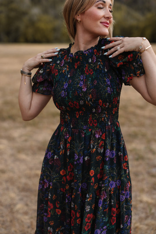A woman with light brown hair stands outdoors on dry grass, smiling as she holds the ruffled sleeves of her Ivy City Co Cordelia Dress in Evergreen Vivid Meadow. The smocked bodice provides a flattering, maternity-friendly fit, and she finishes the look with jewelry on her fingers and wrists.